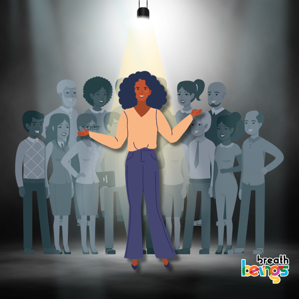 A confident, curly haired, dark skinned stands in the spotlight where a bunch of employees cheer for her in the background. The background is monotone while the woman is highlighted in color wearing a yellow shirt and a grey trouser. She looks confident and has a smile on her face.